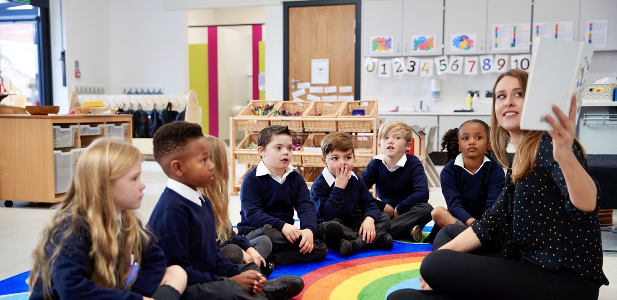 Children in a colourful classroom, sitting in row curving around a female teacher who is holding up a book. 